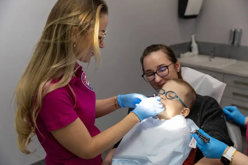 Dentystka bada zęby chłopca, podczas gdy jego mama go wspiera. Zwracają uwagę na zdrowie dziąseł, by zapobiec recesji dziąseł.