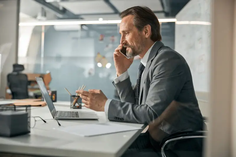 Biznesmen w garniturze rozmawia przez telefon, pracując przy laptopie. To typowy dzień w życiu CEO, co to znaczy być liderem.