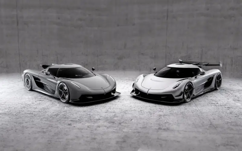 Dwa lśniące Koenigsegg Jesko, jeden szary, drugi srebrny, stoją obok siebie na betonowym tle, gotowe do wyścigu.