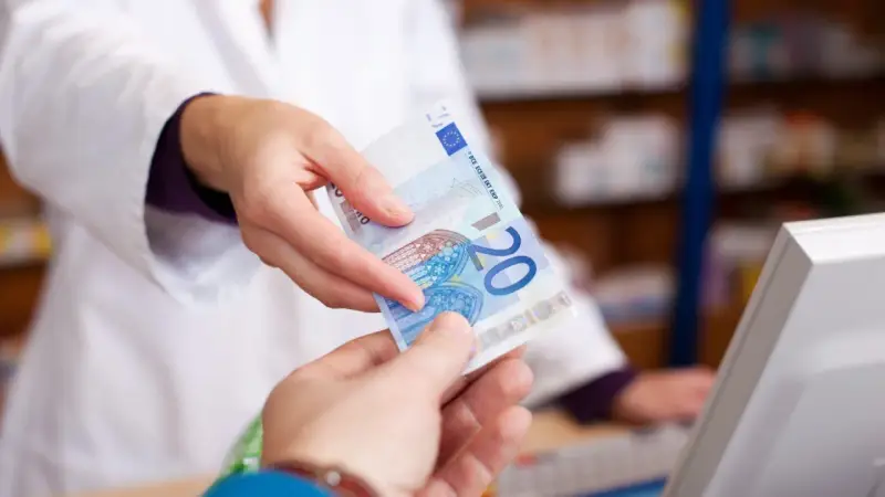 Eine Person in einem weißen Kittel reicht einen 20-Euro-Schein. Möglicherweise zur Bezahlung von Krankengymnastik Kosten.