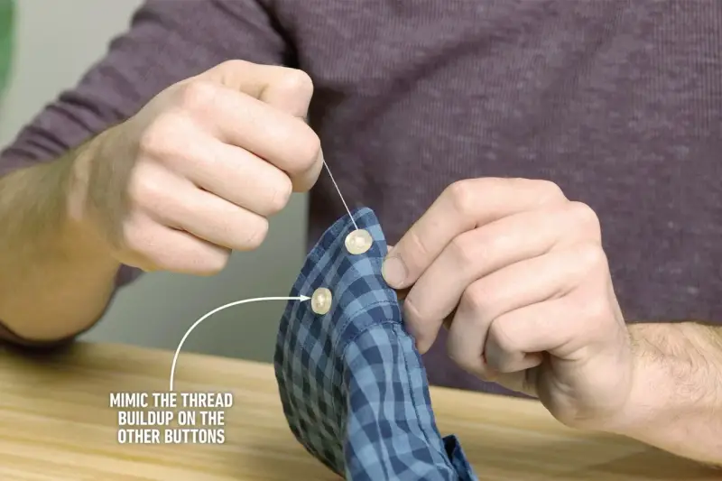 Main tenant une chemise à carreaux bleus, il est en train de coudre un bouton avec une aiguille et du fil blanc.