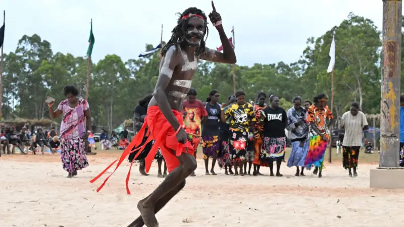 Ein junger Mann mit traditioneller Bemalung und rotem Lendenschurz tanzt. Im Hintergrund stehen Frauen und Männer, die die Zeremonie der Aborigines beobachten.