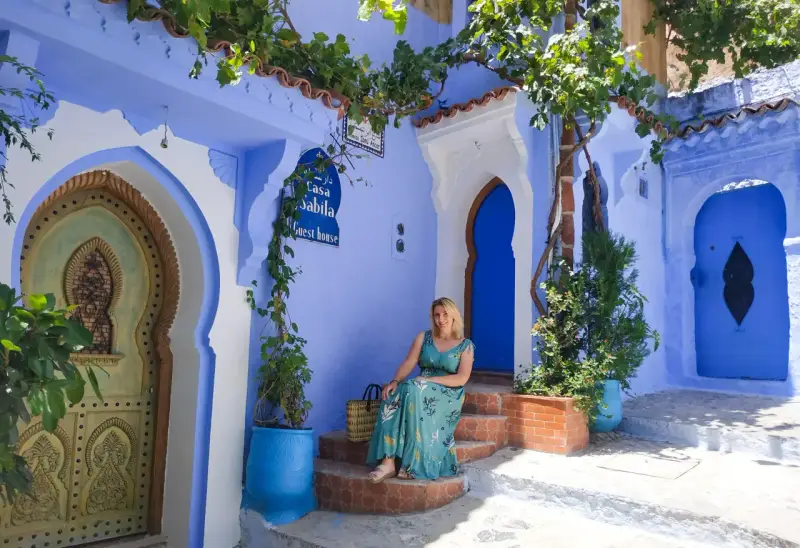 Maj w Maroku to idealna pogoda na zwiedzanie błękitnego miasta Chefchaouen. Kobieta w letniej sukience siedzi na schodach przed Casa Sabila.