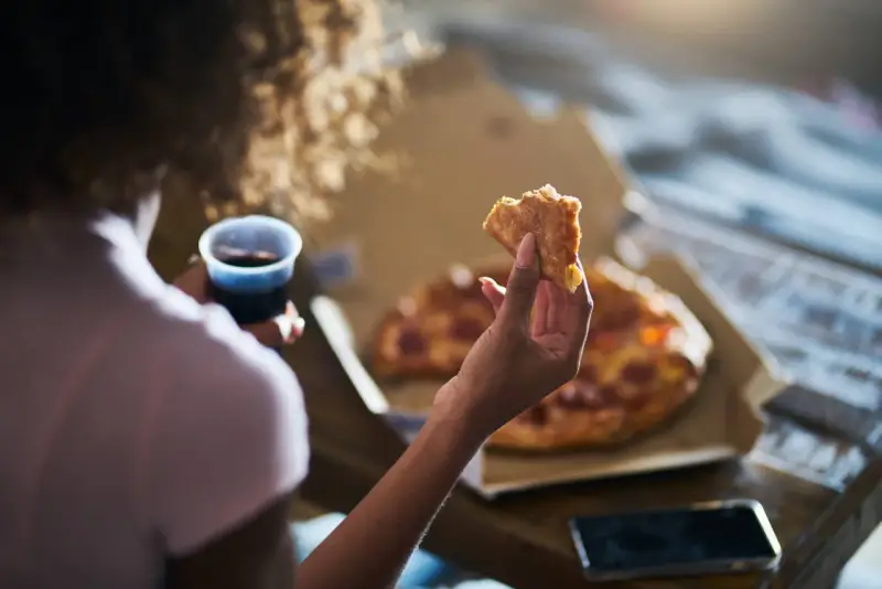 Kobieta trzyma kawałek pizzy i napój, czując głód. Grelina działa, apetyt rośnie.