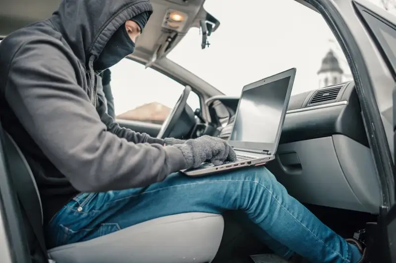 Haker w masce i kapturze próbuje obejść zabezpieczenia keyless go, używając laptopa w samochodzie.