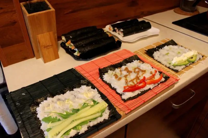 Przygotowanie ryżu do sushi. Na matach leżą arkusze nori z ryżem i dodatkami: ogórkiem, awokado, papryką i sosem.