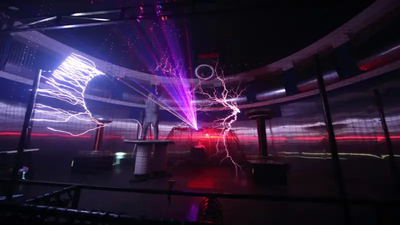 Spektakuläre Blitze zucken in einem **Faradayschen Käfig**, während ein Mann sicher darin steht. Laserstrahlen durchschneiden die Dunkelheit.