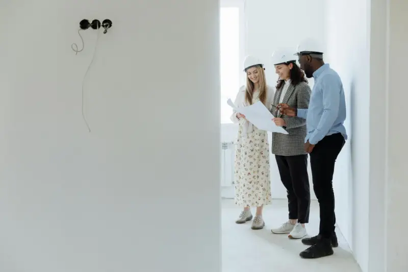 Tres arquitectos con cascos revisan planos en una obra, buscando cómo abaratar costes.