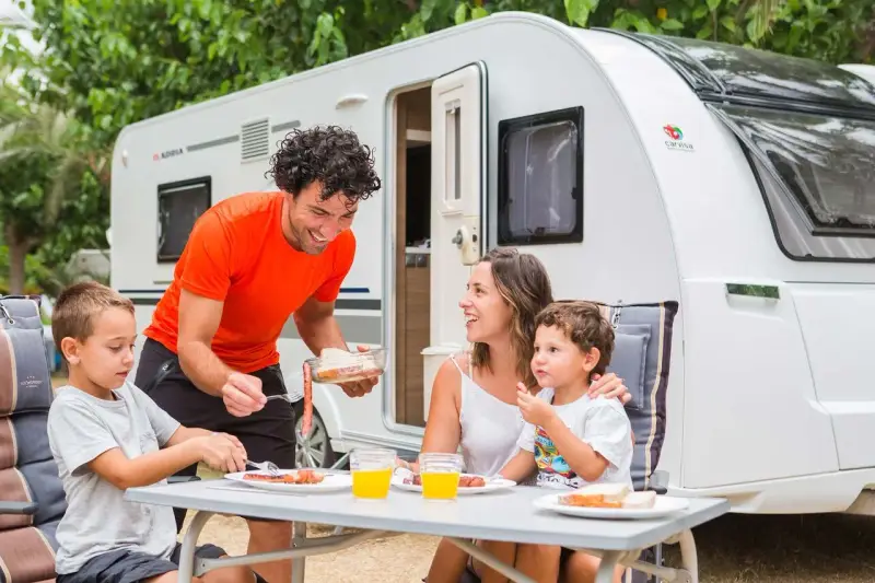 Familia disfrutando de un picnic en campamentos para familias, con una caravana de fondo.