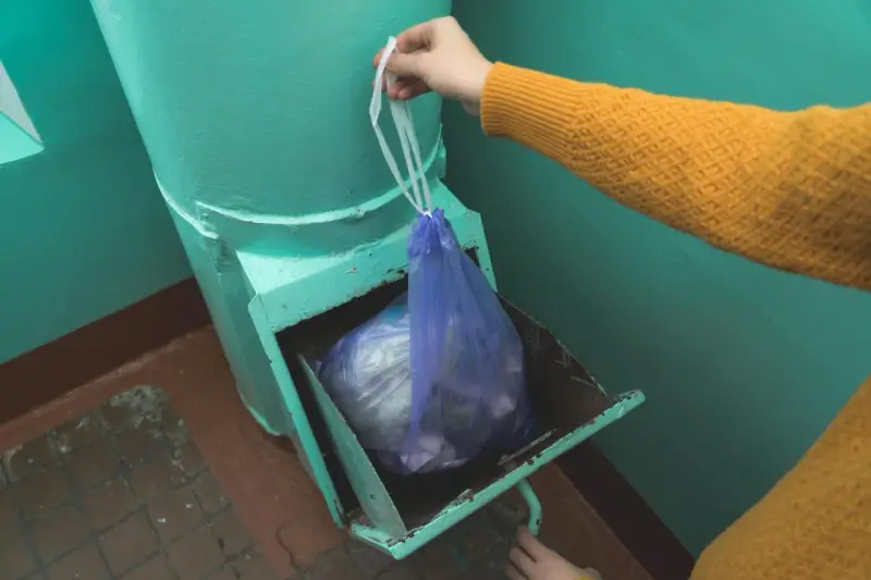 Ręka w żółtym swetrze wkłada worek ze śmieciami do metalowego zsypu w bloku.