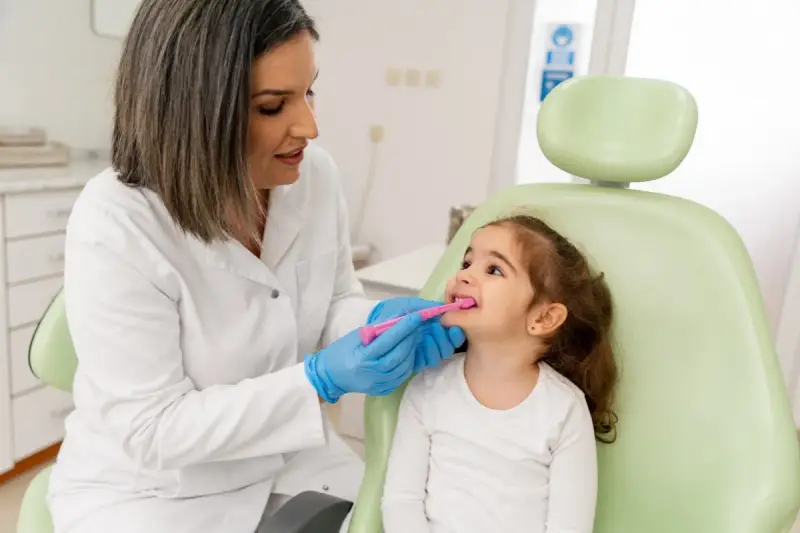 Dentystka w białym fartuchu i niebieskich rękawiczkach pokazuje małej dziewczynce, jak prawidłowo szczotkować zęby. To przykład troski o zdrowie jamy ustnej w ramach pedodoncji.