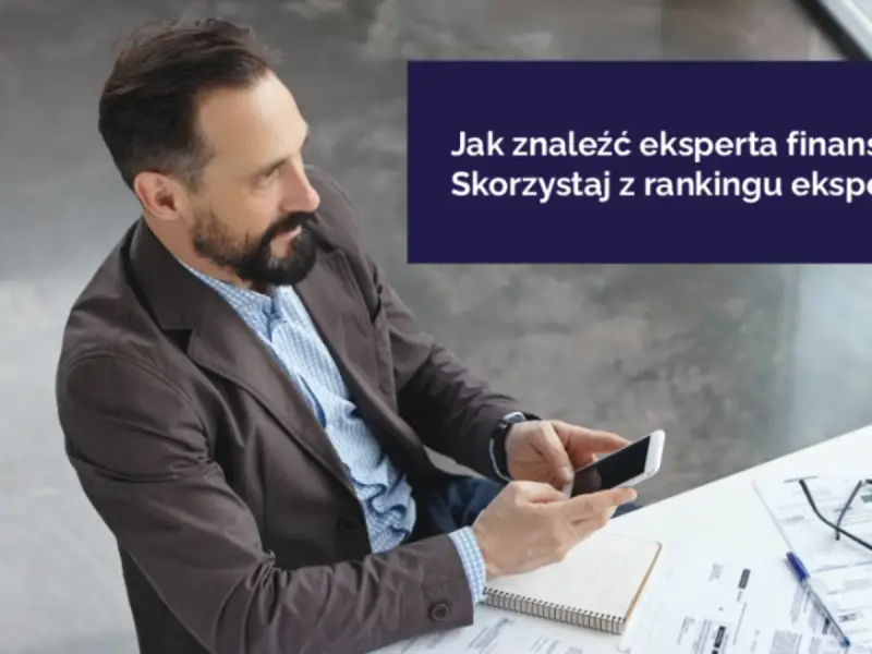 Mężczyzna z brodą trzyma smartfon, siedząc przy biurku z dokumentami.