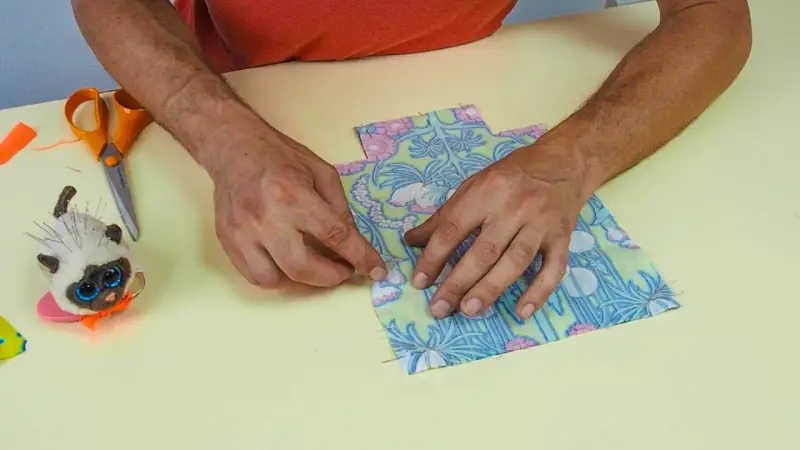 Mains assemblant deux pièces de tissu fleuri, parfait pour une idée couture débutant. Ciseaux et un chat porte-épingles à proximité.
