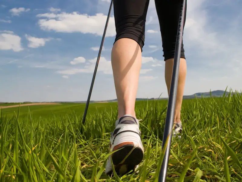 Gambe che praticano nordic walking tecnica su un prato verde sotto un cielo azzurro con nuvole bianche.