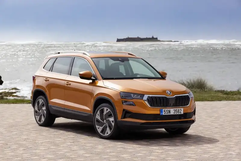 Pomarańczowa Skoda Karoq na tle morza i zamku. Sprawdź aktualny skoda cennik i wybierz swój wymarzony model.