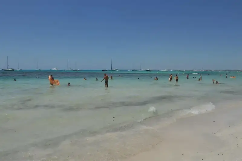Słoneczny dzień na plaży Es Trenc. Ludzie kąpią się w turkusowej wodzie, a na horyzoncie widać żaglówki.