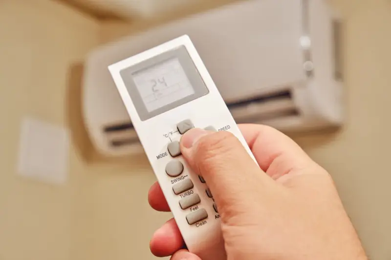 Dłoń trzymająca pilot do klimatyzatora, ustawiająca temperaturę 24°C. Ogrzewanie klimatyzatorem zapewnia komfort.
