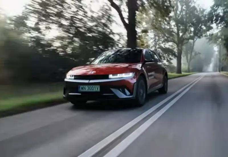 Czerwony Hyundai Ioniq 5 jedzie po wiejskiej drodze wśród drzew.