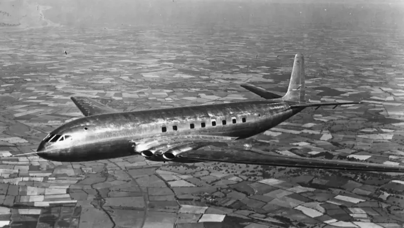 Pierwszy samolot pasażerski, lśniący de Havilland Comet, przelatuje nad patchworkiem pól.