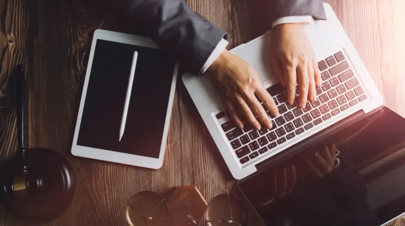 Dłonie na klawiaturze laptopa, obok tablet i młotek sędziowski. Profesjonalne narzędzia do pracy, być może związane z **taxat windykacja**.