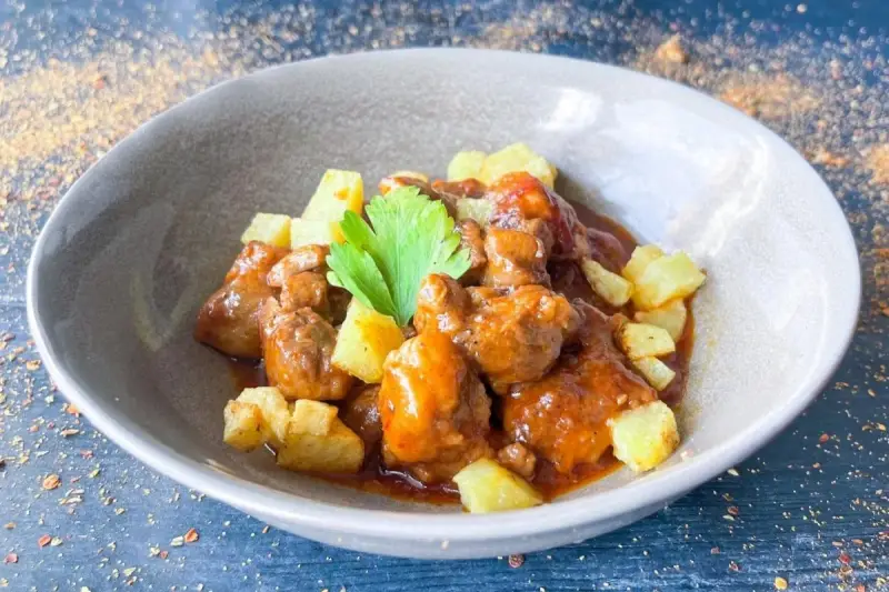 Delicioso cordero al chilindrón en un cuenco gris, acompañado de patatas fritas y una ramita de perejil.
