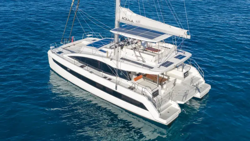 Un catamaran blanc, une véritable bicoque bateau, navigue sur une mer bleue. Ses voiles sont partiellement déployées, et des panneaux solaires brillent sur son toit.