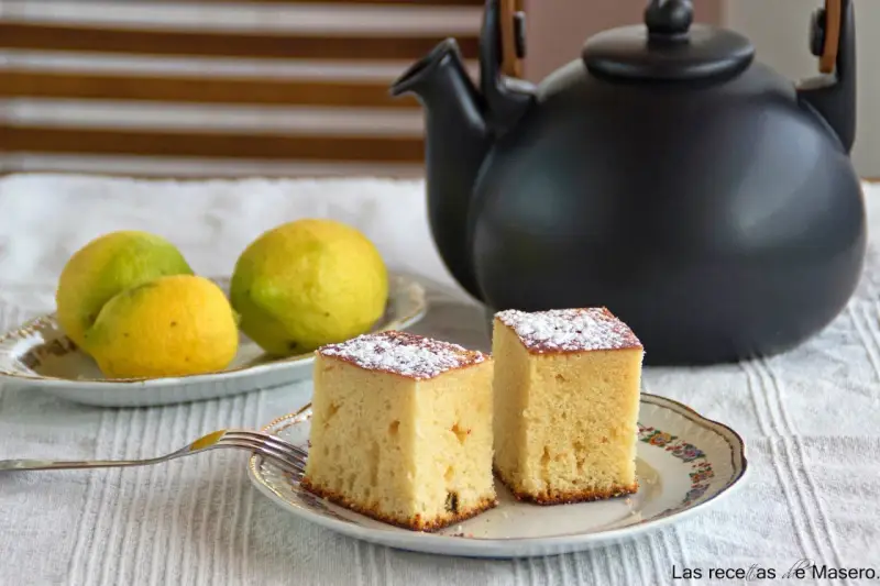 Dos porciones de bizcocho de limón espolvoreadas con azúcar glas, junto a limones frescos y una tetera negra.