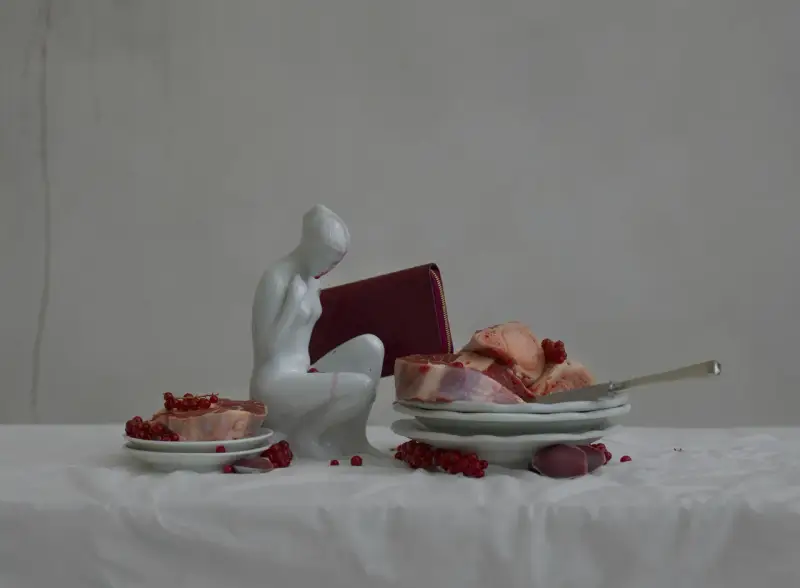 A white porcelain figure contemplates raw meat and red currants, a scene reminiscent of Evelyn Bencicova's still lifes.