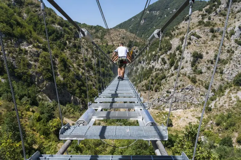 Un ponte tibetano in Puglia attraversa una gola verdeggiante, con persone che camminano su passerelle metalliche sospese tra montagne.