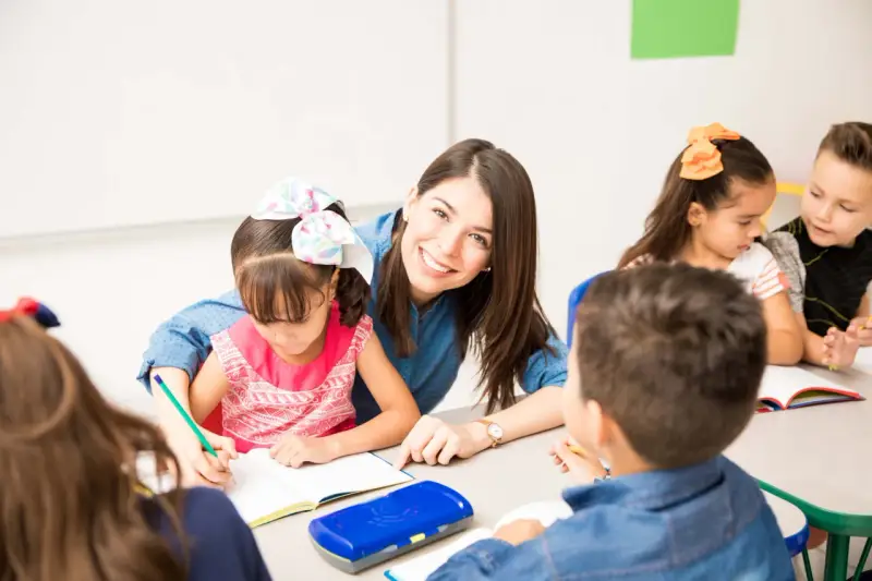 Maestra sonriente ayuda a una niña a escribir. Un aula llena de niños y frases bonitas para una profesora.