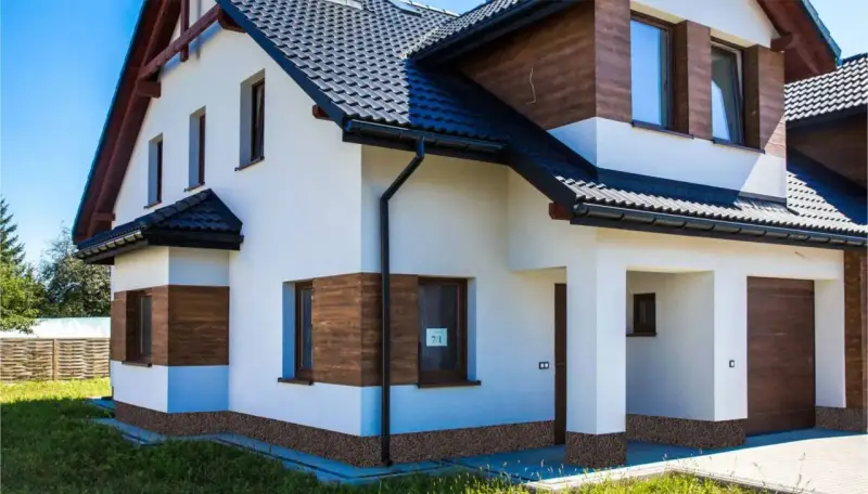 Nowoczesny dom z białym tynkiem i drewnianymi akcentami. Widoczny ciemny dach, okna i drzwi. Dookoła domu biegnie ozdobny cokół.