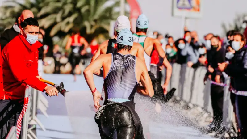 Zawodnicy triathlonu olimpijskiego biegną, a strażak polewa ich wodą z węża, by ich ochłodzić. Widownia w maskach obserwuje zza barierek.