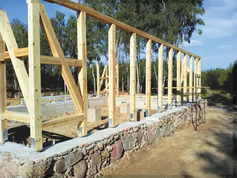 Drewniana konstrukcja domu powstaje na solidnym fundamencie z kamienia. Widać betonowe słupy i kamienny murek.
