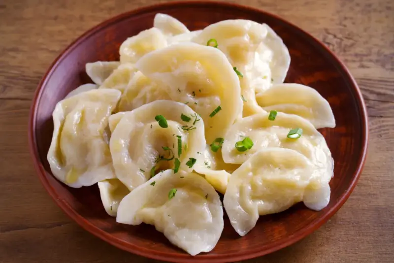 Pyszne pierogi z tradycyjnym farszem do uszek, posypane szczypiorkiem, na glinianym talerzu.