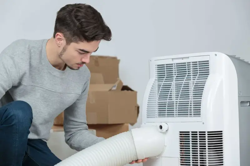 Un jeune homme installe un climatiseur mobile 40m2. Des cartons sont empilés derrière lui.