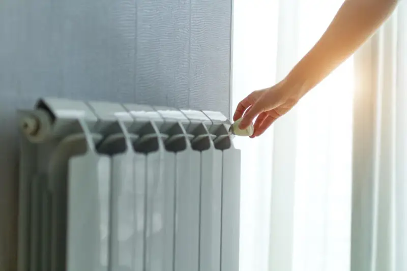 Dłoń reguluje termostat na grzejniku, pokazując różne rodzaje kaloryferów i kontrolę temperatury w domu.
