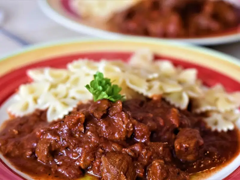 Gulasz z indyka z warzywami, podany z makaronem w kształcie motylków, ozdobiony natką pietruszki. Pyszne danie na talerzu.