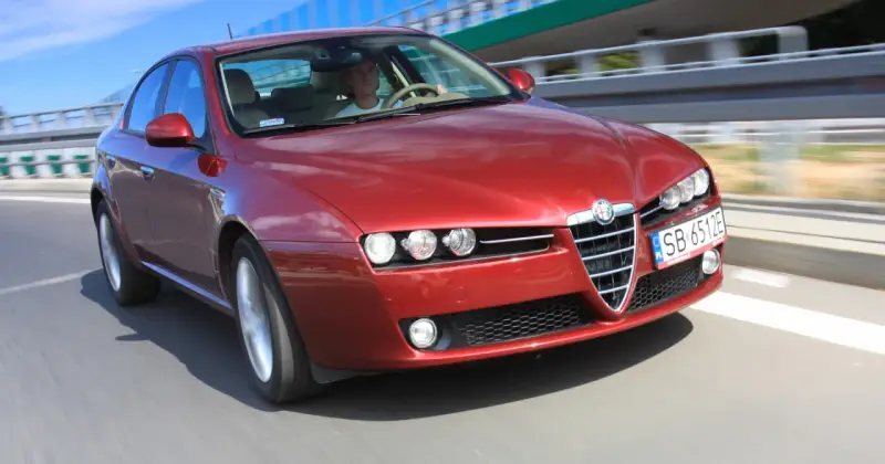Czerwone Alfa Romeo 159 na drodze. Kierowca w białej koszulce, pozytywne opinie o tym modelu potwierdzają jego sportowy charakter.