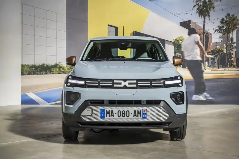 Jasnoszary dacia elektryk z miedzianymi lusterkami. Z przodu logo Dacia i tablica rejestracyjna MA-080-AM 92.