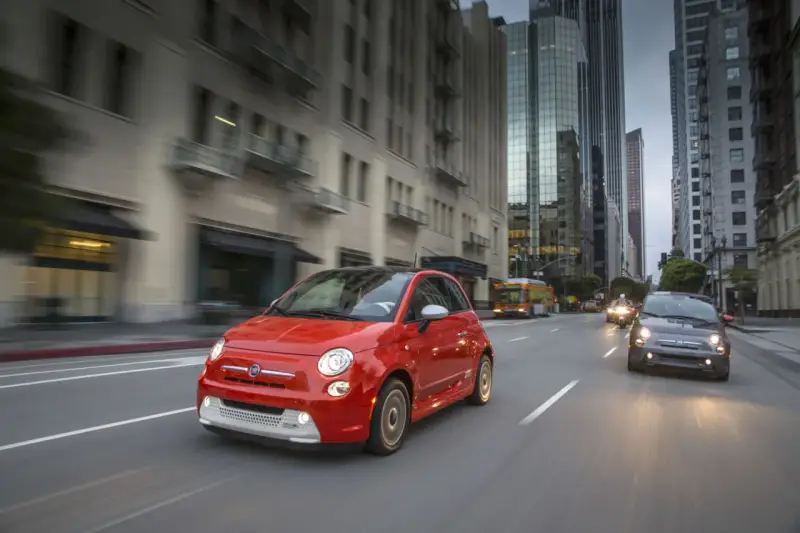 Czerwony Fiat 500e jedzie ulicą w mieście, w tle wieżowce i autobus.