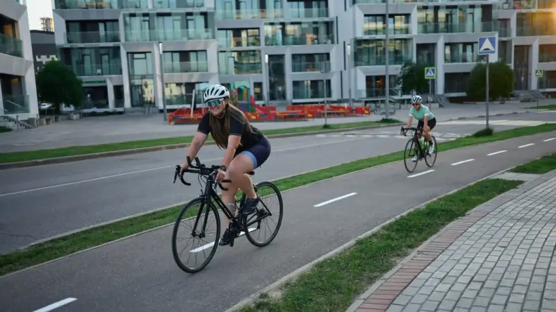 Zwei Rennradfahrerinnen genießen eine Fahrt auf einem Radweg vor modernen Gebäuden.