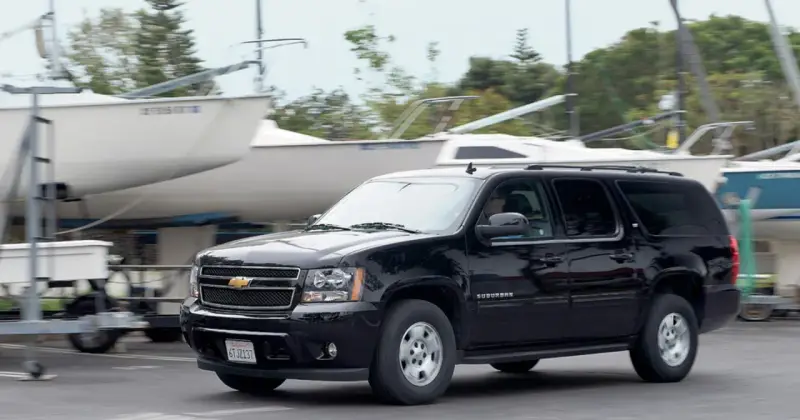 Czarny Chevrolet Suburban jedzie obok przyczep z łodziami, w tle drzewa.