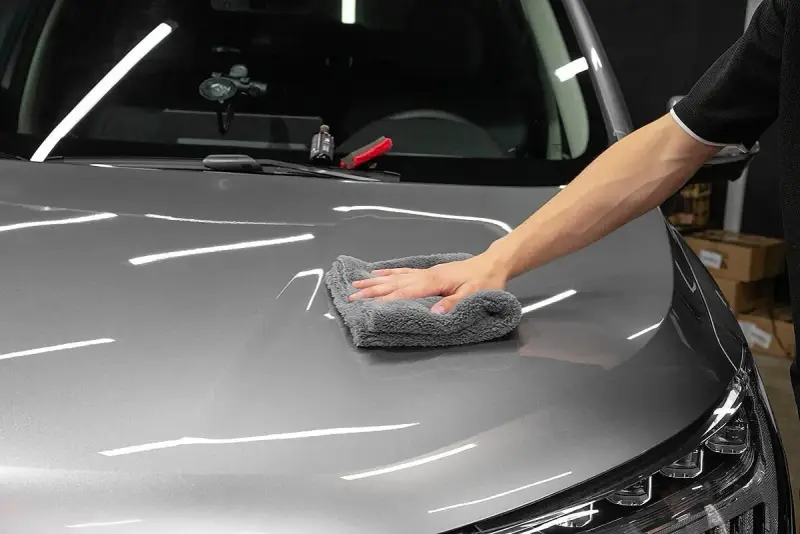 A hand buffs a car's hood with a microfiber cloth, applying a ceramic wax for a deep shine.