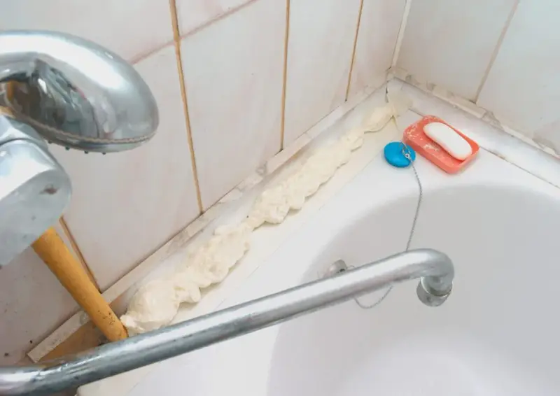 Mousse expansive appliquée pour nettoyer joint plastique porte douche. Savon et bouchon de bain à proximité.