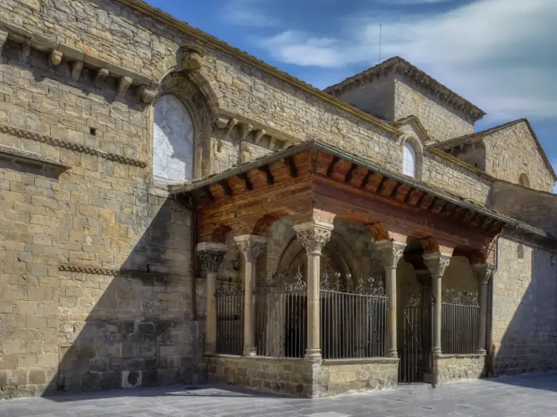 La **catedral de Jaca**, con su imponente arquitectura románica, se alza bajo un cielo azul. Un pórtico de madera y piedra protege la entrada.
