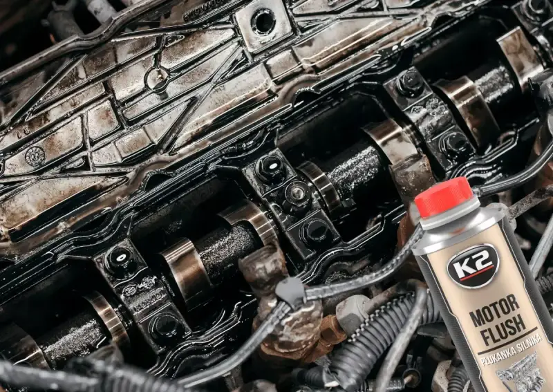 Silnik z widocznymi elementami rozrządu, pokryty czarnym osadem. Obok butelka płukanki K2 Motor Flush, która pomoże rozwiązać problem niskie ciśnienie oleju na rozgrzanym silniku.
