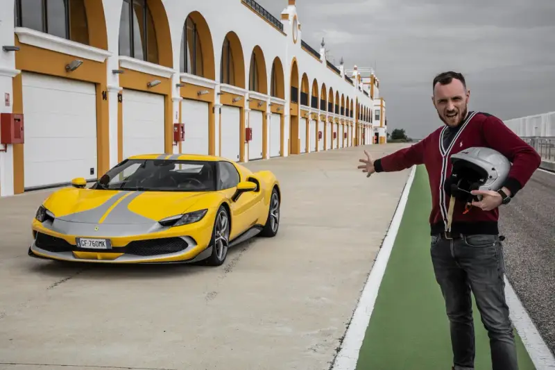 Żółte Ferrari 296 GTB z szarymi pasami na torze wyścigowym. Obok stoi mężczyzna z kaskiem, zapraszając do przejażdżki.