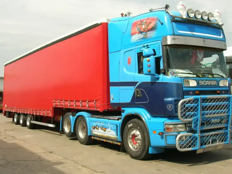 Naczepa jumbo w jaskrawym kolorze czerwonym, ciągnięta przez niebieską Scanię z malowidłami.