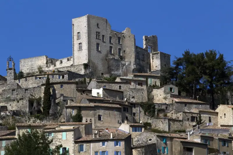 Vue d'un village perché dominé par les ruines imposantes du **château du marquis de Sade**, sous un ciel bleu éclatant.