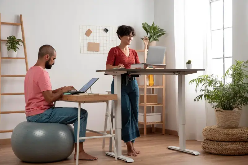 Un homme travaille sur son ordinateur, assis sur un ballon de gym, tandis qu'une femme travaille debout à un bureau réglable. Un espace de travail moderne et dynamique.